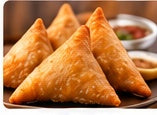 Two golden-brown samosas on a dark plate with chutneys in the background